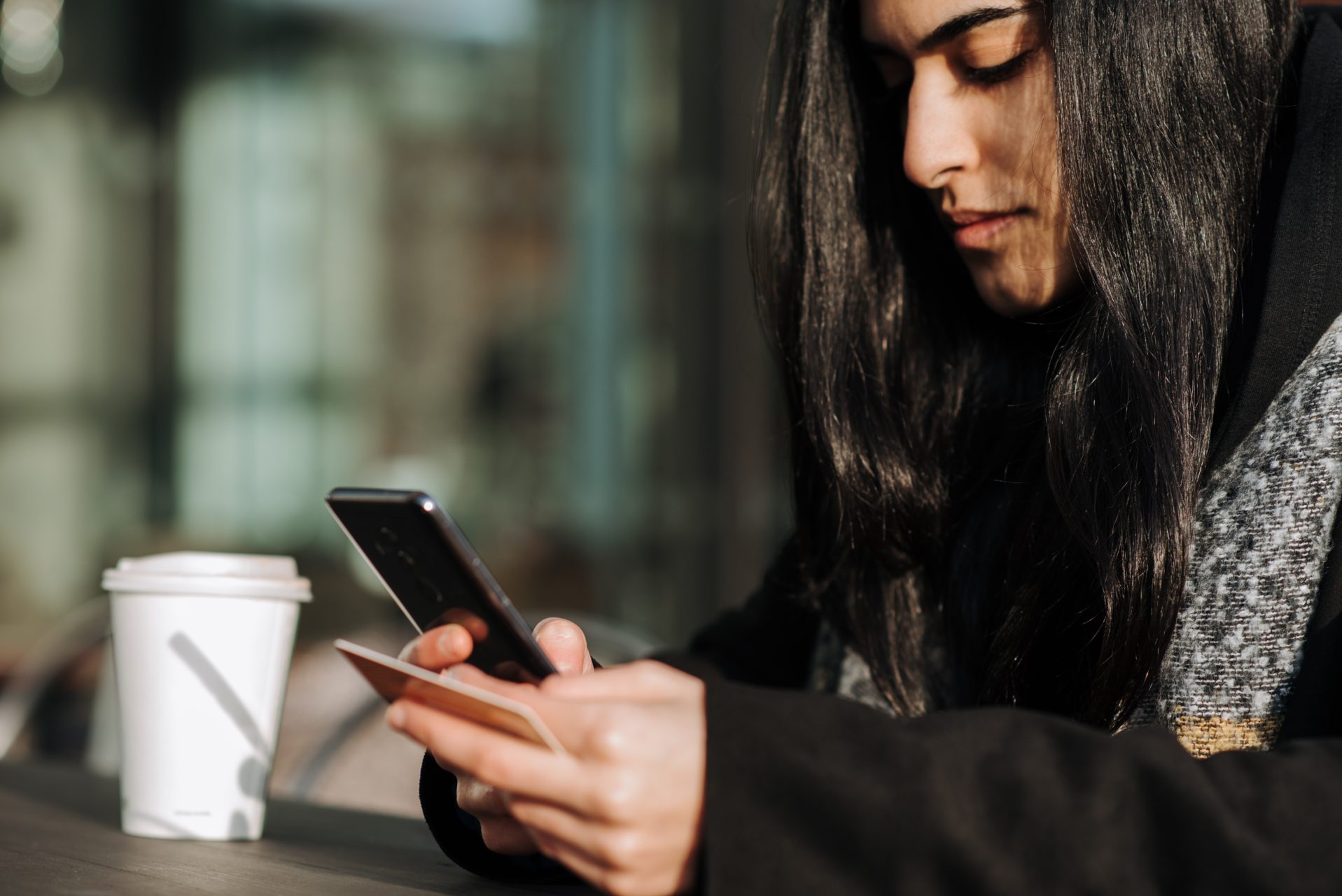 femme assise avec un café devant elle est entrain d'utiliser son smartphone et sa carte de crédit