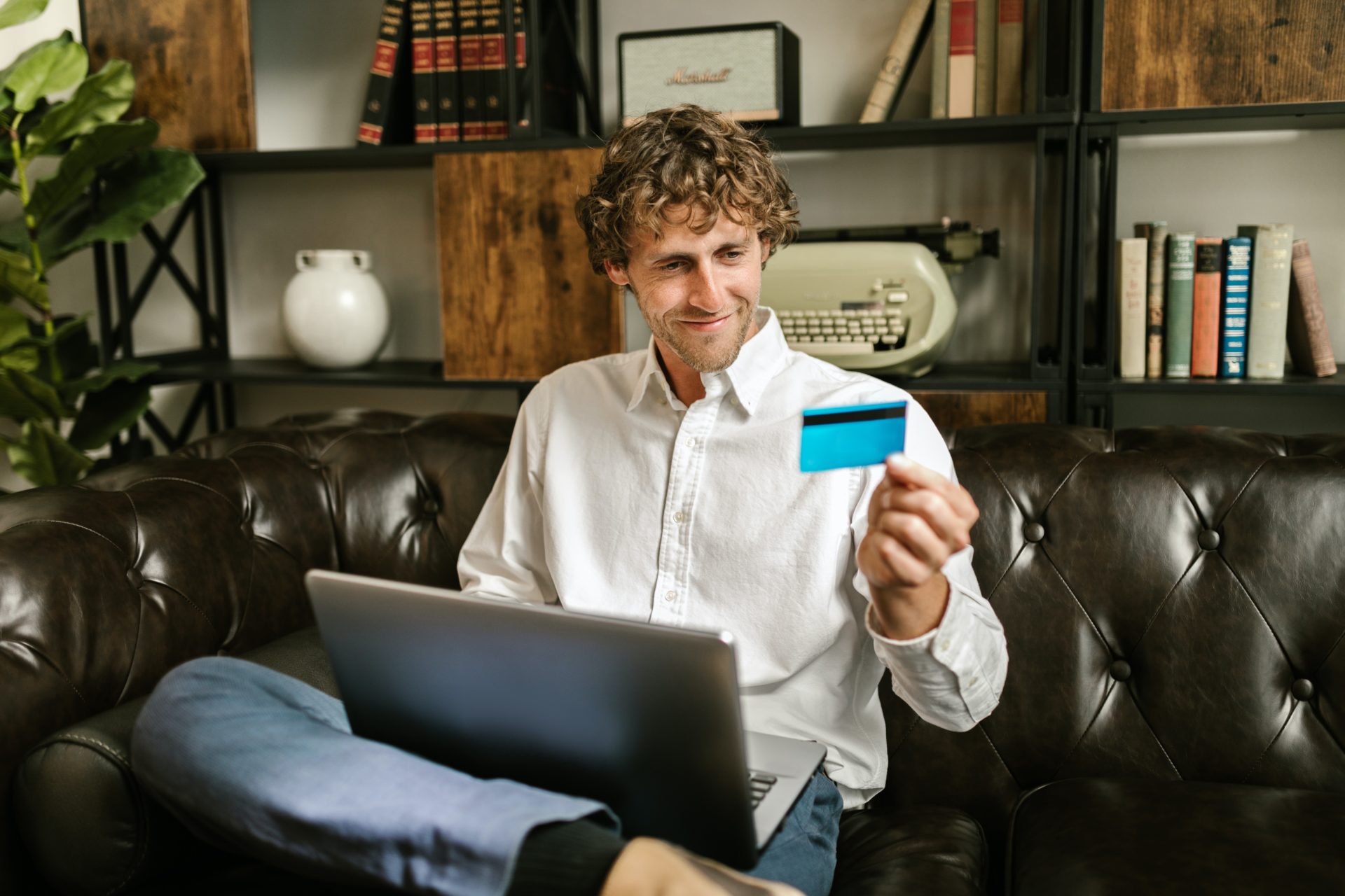 homme avec un ordinateur portable sur ses genoux est assis et sourit en regardant sa carte de banque qju'il tient dans sa main gauche