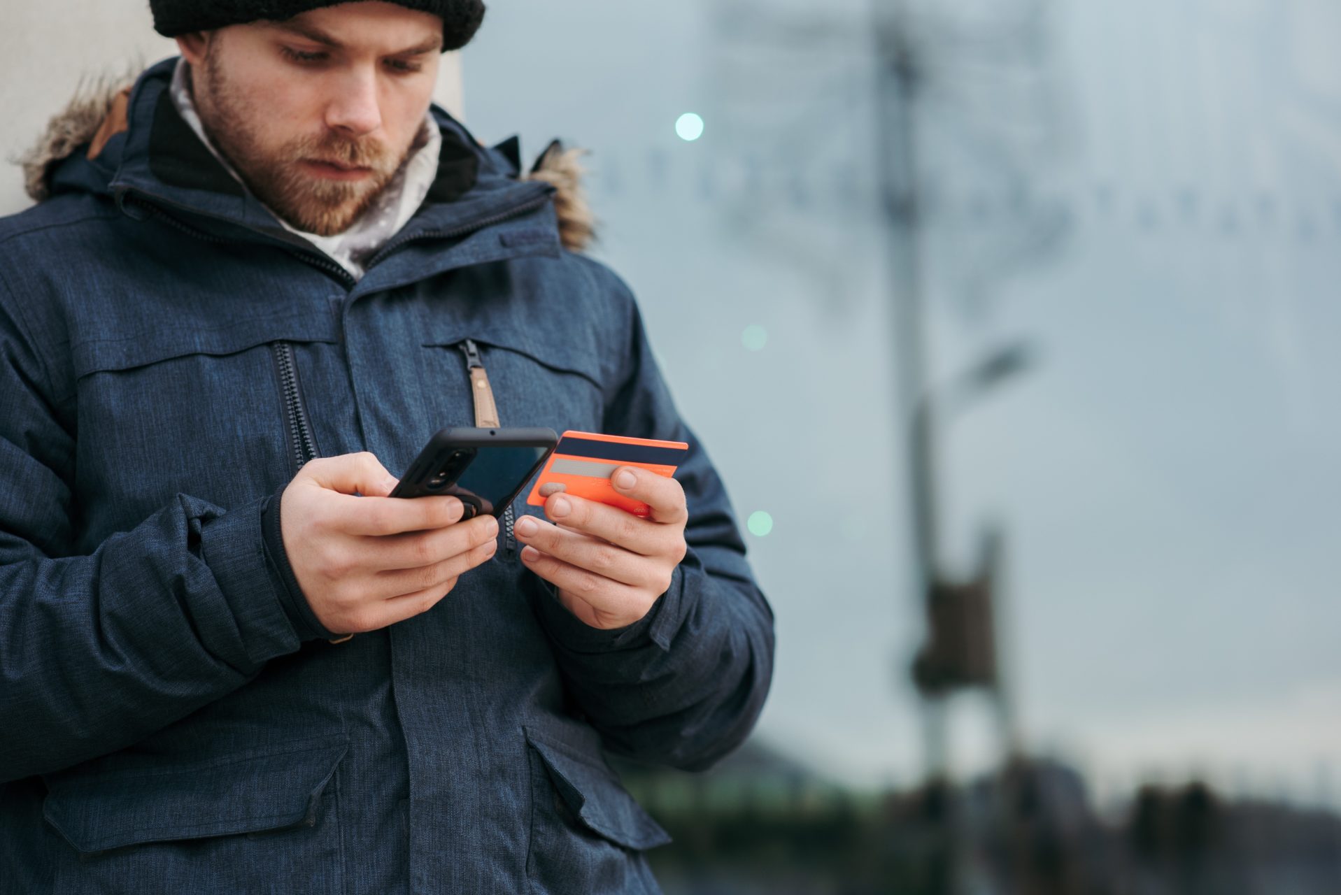 homme en manteau bleu tient dans ses mains son smartphone et sa carte de crédit