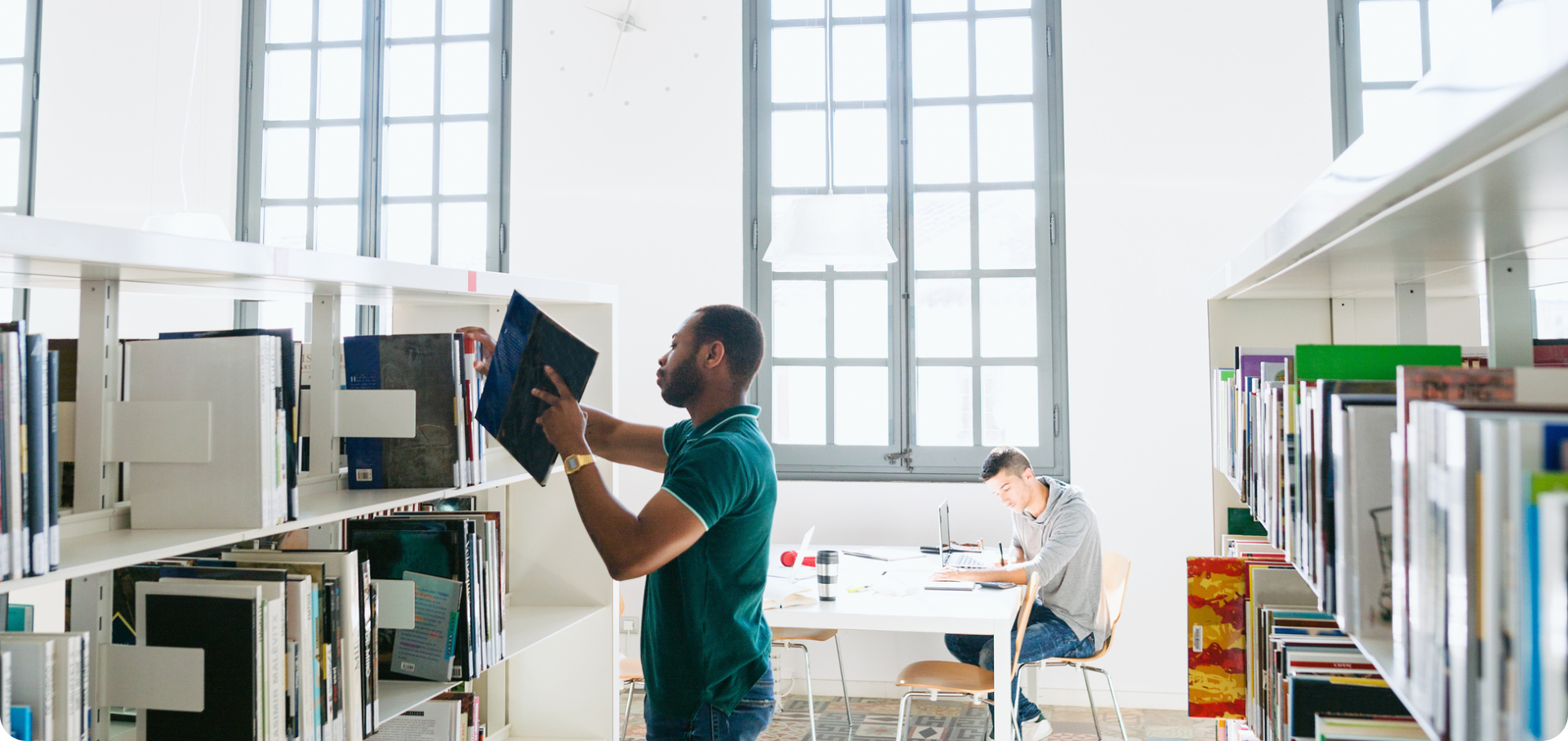 Image on the business expenses page; Two men in an office or library. One is sat working at a desk, while the other is returning a book to the shelf in a library.