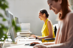 Representing Soldo's Tailored Solutions: Two women at their desks, smiling and typing on their computers. Both women are on the right side of the image, facing left. One is in a yellow blouse, the other is in pink. There is a stack of papers and a plant on the desk.